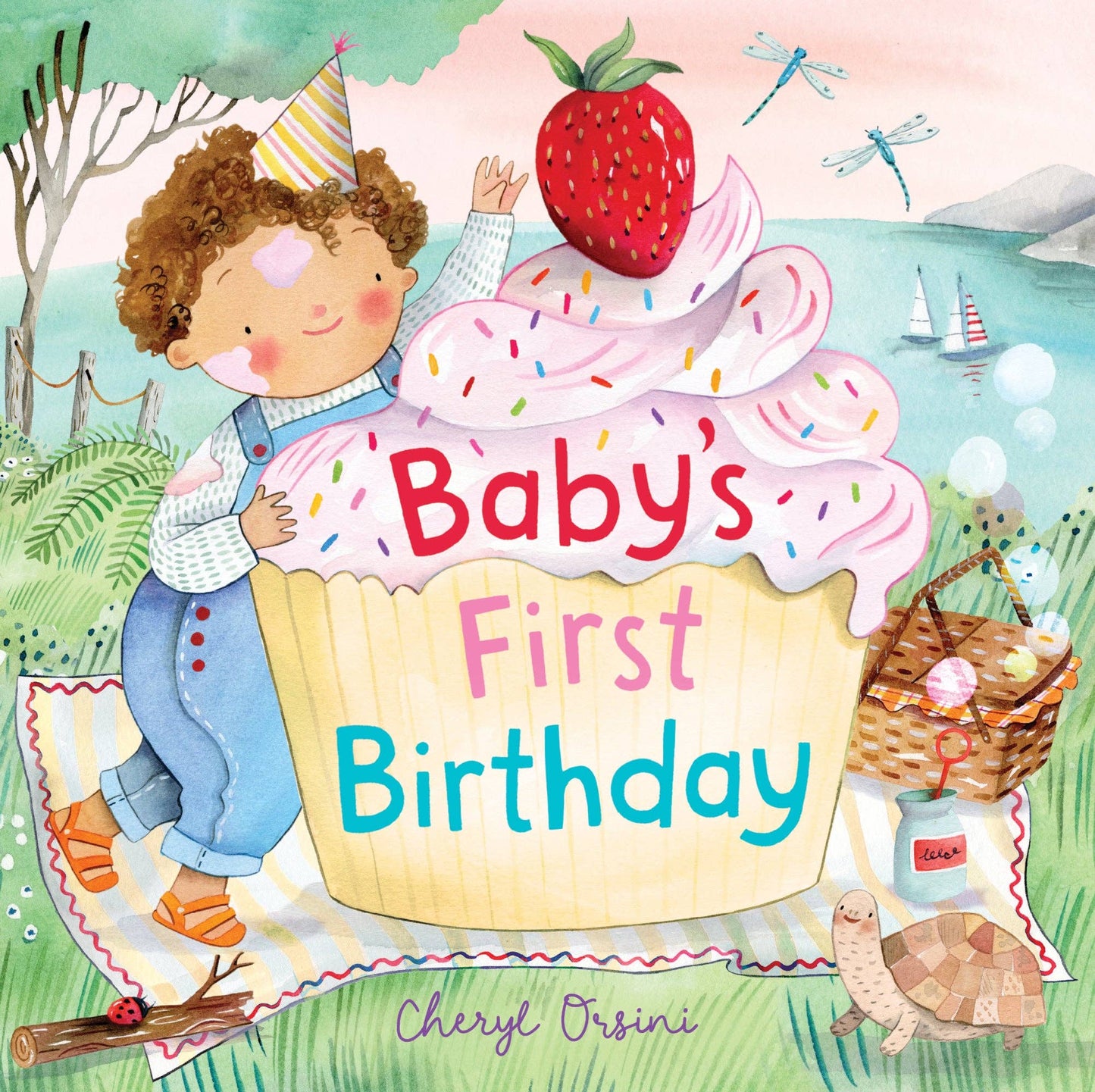 Illustrated children's book cover titled "Baby's First Birthday" by Cheryl Orsini, showing a baby peeking from behind a giant strawberry cupcake on a picnic blanket by the water.
